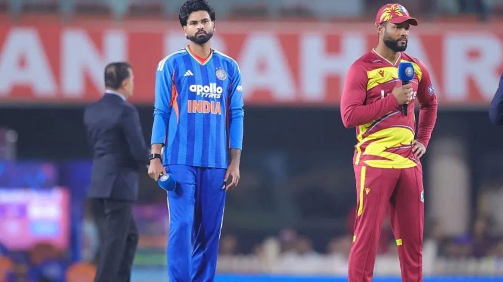 India and West Indies players stand at the toss in their team kits, the India player in a blue jersey and the West Indies player in a maroon and yellow kit holding a microphone, with a broadcaster beside them