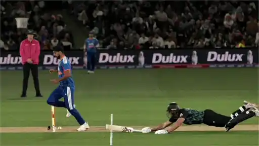 A bowler in India’s blue kit stands beside broken stumps with the ball at his feet, while a New Zealand batter in a black kit dives full-length with his bat lifted just short of the crease, as an umpire in pink watches