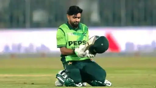 Pakistan cricketer in a green jersey kneeling on the field and looking down at his helmet and bat, with the stadium blurred in the background.