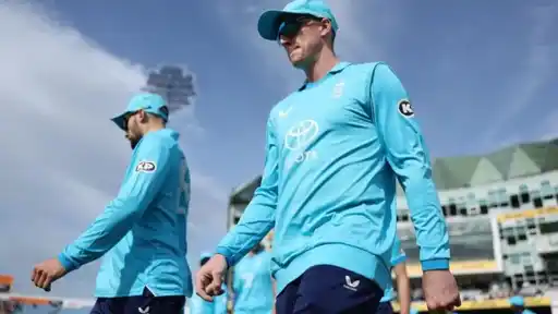 Two England players in light blue kit and caps walking across a ground, with a stadium stand and cloudy sky behind them.