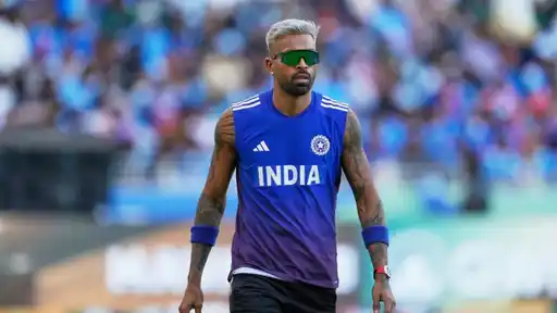 Cricketer in a blue sleeveless India training jersey, green mirrored sunglasses and blue wristbands walks across a ground with a blurred crowd behind him