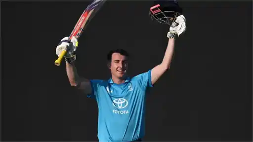 A smiling cricketer in a light blue England kit raises his bat in one hand and his helmet in the other against a dark background