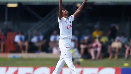 Hasan Mahmud in Bangladesh’s white Test kit stretches both arms skyward, looking up in celebration, with spectators seated on a grass bank behind him