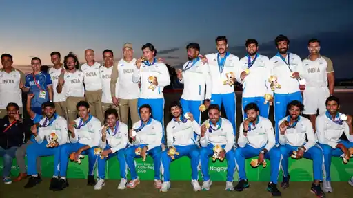 India men’s cricket squad and support staff pose in India kit with gold medals and mascot toys on the podium at the Aichi-Nagoya 2026 Asian Games, after retaining their title.