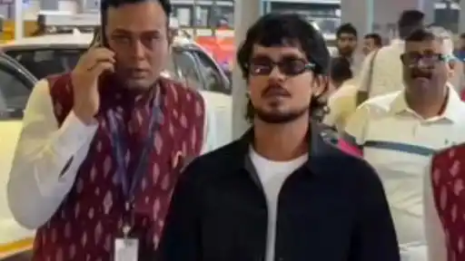 Young man in tinted sunglasses, a black jacket and white t-shirt walks through a crowded area beside a man in a red patterned vest on a phone call, ahead of the IND vs WI T20Is