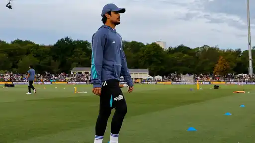 Ishan Kishan in a blue India training hoodie and cap stands on the outfield with fans in the stands behind him, before India’s 3rd T20I against New Zealand at Wellington