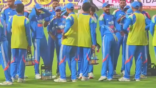 India players in blue kit huddle on the field as one laughs and another bends forward mid-move, while substitutes in yellow bibs carry drinks during the IND vs WI 3rd ODI