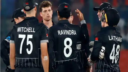 New Zealand players in black ODI kit gather in a huddle during a match, with the names Mitchell, Ravindra and Latham visible on the backs of their shirts.