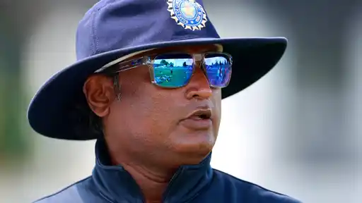 Close-up of a man in a navy BCCI wide-brimmed hat, mirrored sunglasses and a navy zip jacket, with a cricket field reflected in the lenses