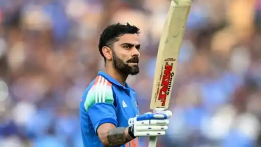Virat Kohli in India’s blue ODI jersey raising his bat to acknowledge the crowd, with a blurred stadium crowd behind him.